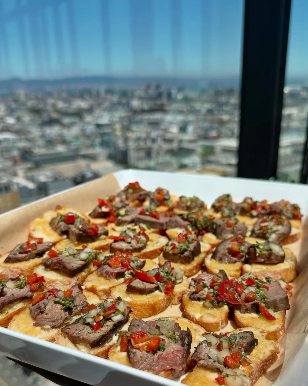 Tray of steak crostini with chimichurri at a high-rise event, the San Francisco skyline through the window