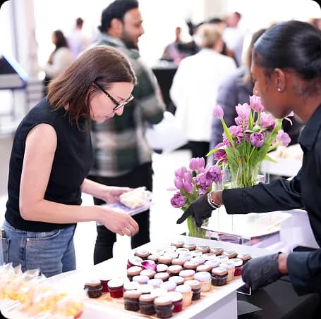 Guests choosing logo-topped cupcakes from an office dessert table as a Pinx server tends the display