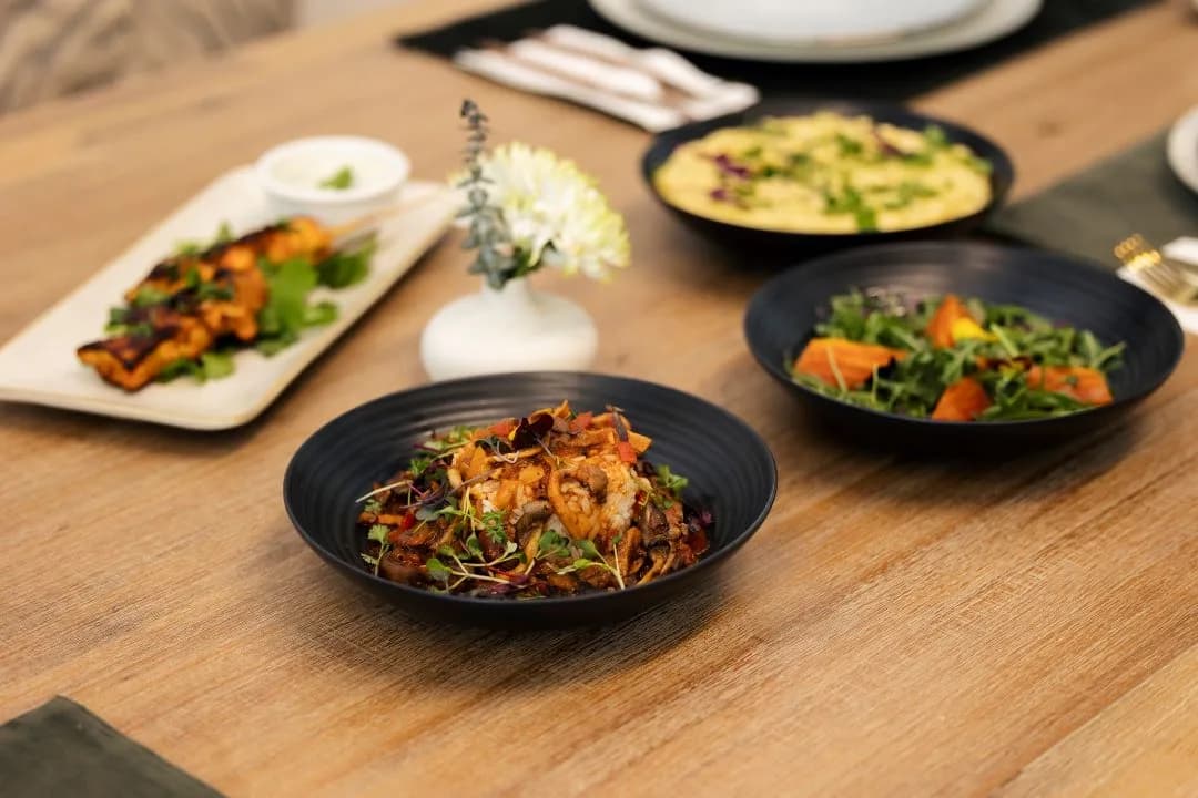 Black bowls of rice with mushrooms, arugula and roasted squash salad, pasta and a skewer platter on a wood table