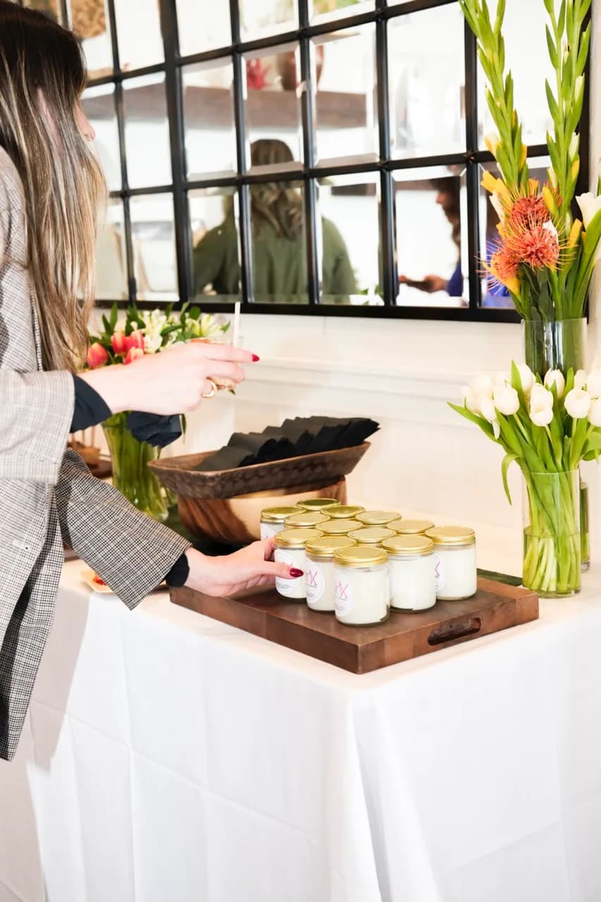 A guest taking a jarred Pinx favor from a wooden tray on a white-linen table set with tulips and protea
